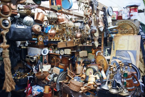 A densely packed outdoor display of miscellaneous household and decorative items, including copper and brass pots, lamps, and vases, arranged on shelves and directly on the ground. The objects vary in size, shape, and finish, with some polished and others aged or tarnished, exhibiting warm metallic tones and textured surfaces. The scene includes small woodenware, framed artworks, and various types of crockery, with some items hanging on or leaning against the cluttered backdrop, which appears to be a market or junk stall in a busy courtyard or street setting. Natural daylight illuminates the scene, highlighting the contrasts between shiny metals and aged finishes, creating a vibrant but cluttered appearance indicative of a sale or clearance of second-hand items. This image relates to waste management and rubbish removal services offered by Commercial Waste Barbican, situated in or near the City of London, supporting sustainable disposal and clearance of household or business waste in central London.