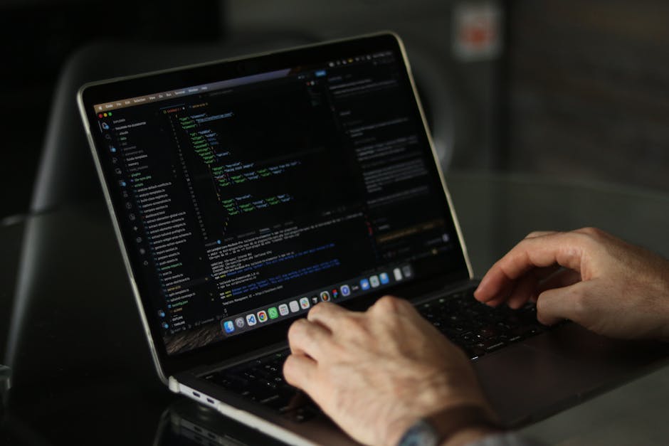 A person working on a silver laptop with a black keyboard, positioned on a dark-colored surface, displaying a coding or programming interface on the screen, which contains multiple lines of colorful c