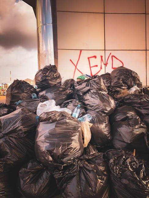 A large pile of black plastic rubbish bags filled with waste is stacked against a building with a tiled exterior wall. Some bags are slightly torn or bulging, revealing glimpses of waste inside, inclu