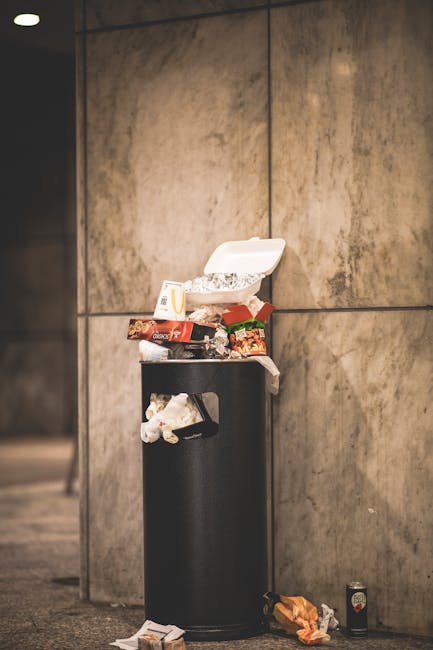 A large pile of black plastic rubbish bags filled with waste is stacked against a building with a tiled exterior wall. Some bags are slightly torn or bulging, revealing glimpses of waste inside, inclu